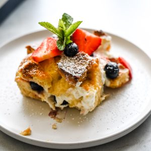A slice of Berries and Cream French Toast Bake topped with fresh strawberries, blueberries, a sprig of mint, and a dusting of powdered sugar sits on a white plate.