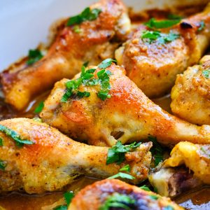 Close-up of cooked Honey Mustard Baked Chicken Drumsticks garnished with fresh parsley in a rich, golden-brown sauce. The chicken is seasoned and has a crispy, golden skin, surrounded by a flavorful broth.