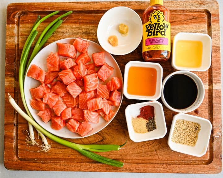 Overhead view of cubed salmon and small bowls of sesame oil, honey, soy sauce, garlic, ginger, and seasonings arranged on a wooden board for Honey Baked Salmon Bites.