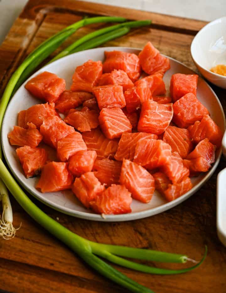 Raw cubed salmon in a bowl with sliced green onions and small bowls of honey, soy sauce, sesame oil, garlic, ginger, and spices arranged on a wooden board.