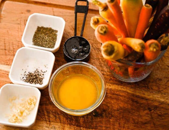 Ingredients for honey butter roasted carrots arranged on a wooden surface, including fresh carrots, melted butter, honey, herbs, and seasonings.
