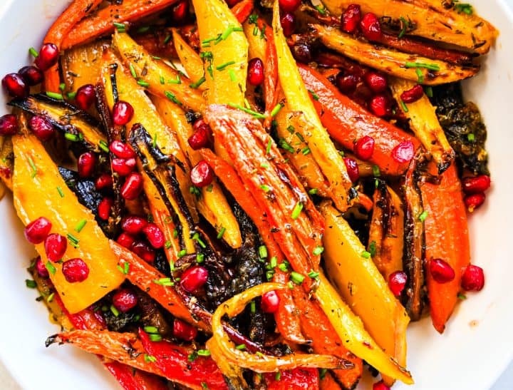 Honey butter roasted rainbow carrots with golden edges, pomegranate seeds, and fresh chives served in a white bowl.